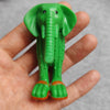 A hand holds a green wooden elephant stick doll figure with orange feet.