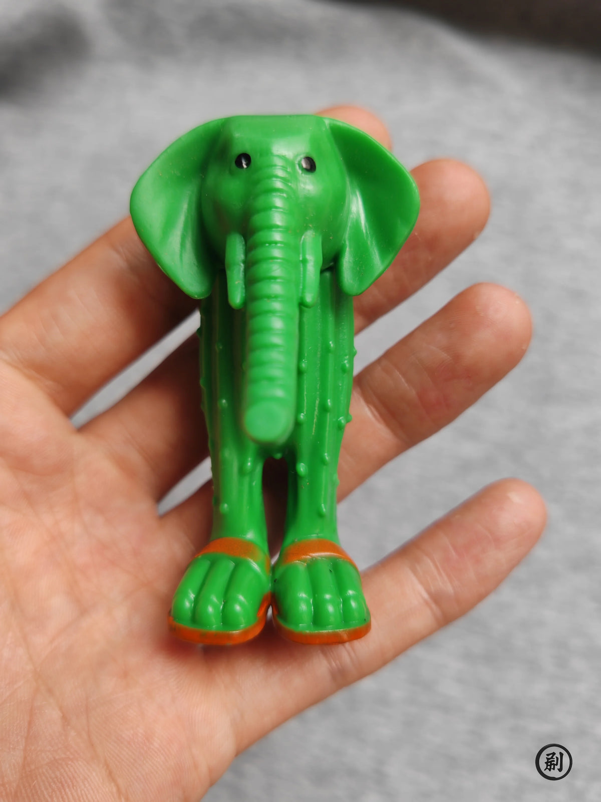 A hand holds a green wooden elephant stick doll figure with orange feet.