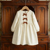 A cream-colored girls' long sleeve A-line dress with brown bow details displayed on a wooden cabinet.