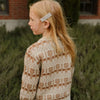 A blonde girl wearing a beige knit sweater and light colored pants outdoors.