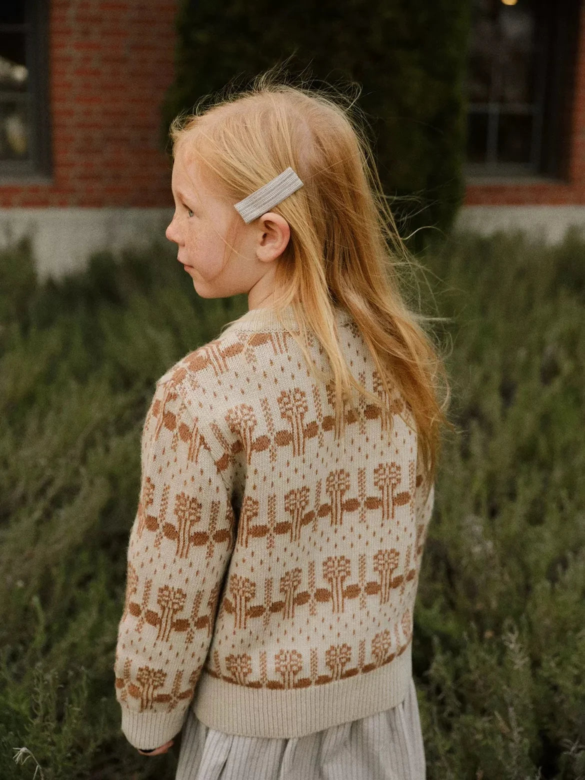 A blonde girl wearing a beige knit sweater and light colored pants outdoors.