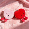 Two plush hearts, one pink and one red, resting on a fluffy pink background.