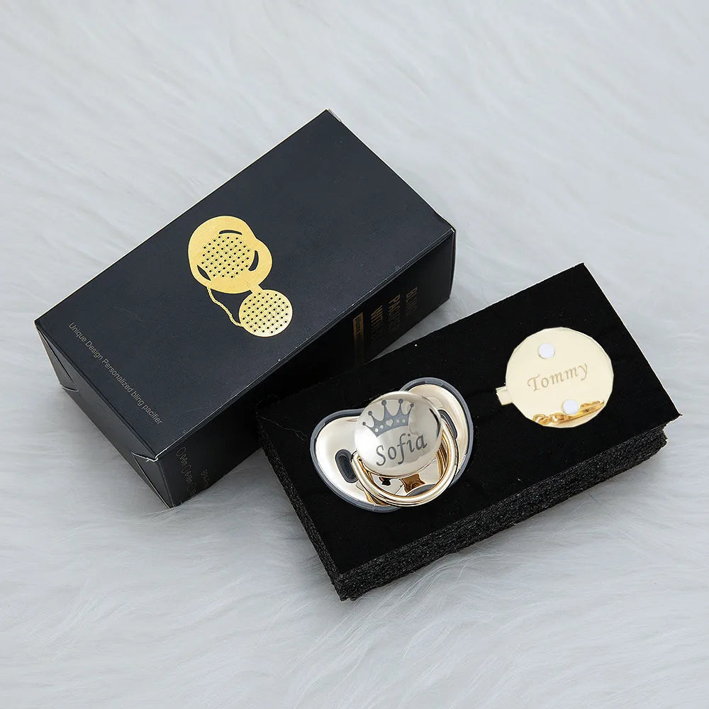 A black box containing a rose gold rhinestone pacifier and clip set with a gold charm.