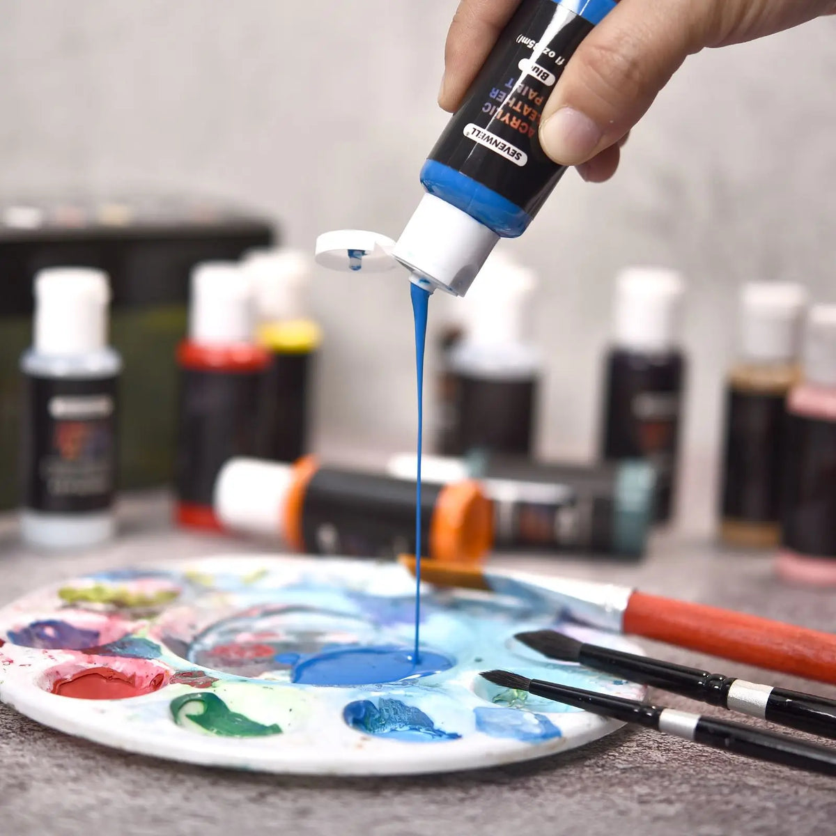 A hand is pouring blue acrylic paint onto a palette of various colored paints for DIY projects.