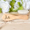 A light wooden baby brush and comb set with the name Julie engraved on the handle, resting on a beige cloth.