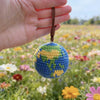 A handcrafted crochet earth keychain charm with blue and yellow yarn is held in a hand against a blurred field of colorful wi