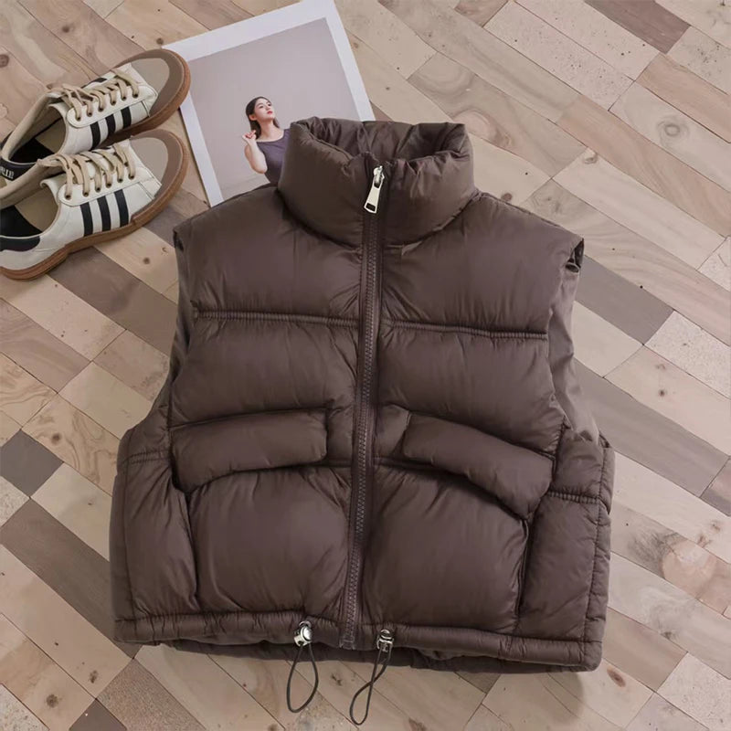 A brown padded down vest is displayed on a wooden floor with a person standing in the background.
