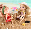 A blonde doll with a straw hat is seated on a wooden chair in the sand next to a table with colorful desserts.