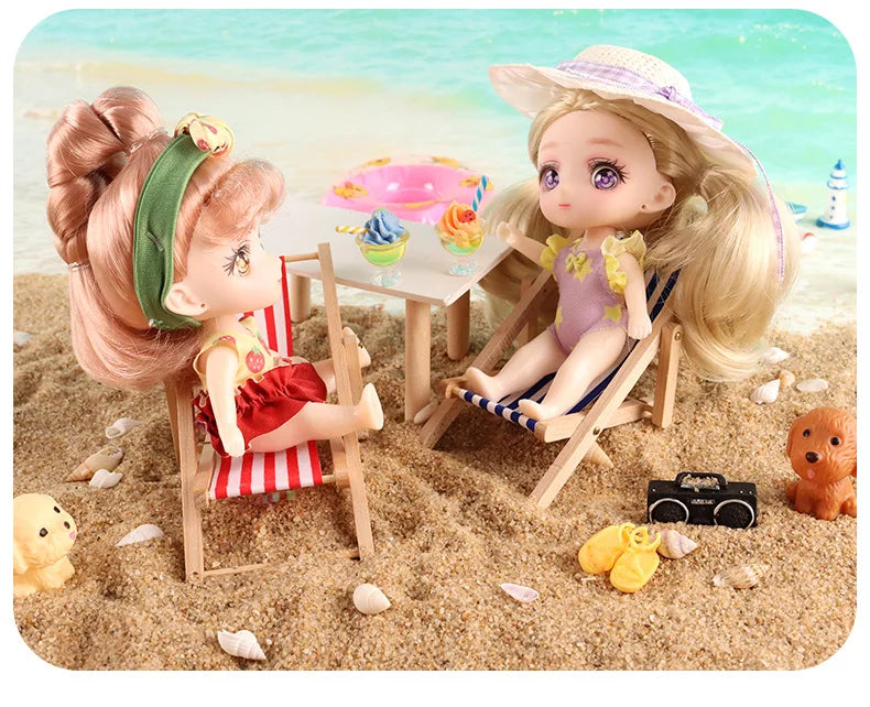 A blonde doll with a straw hat is seated on a wooden chair in the sand next to a table with colorful desserts.
