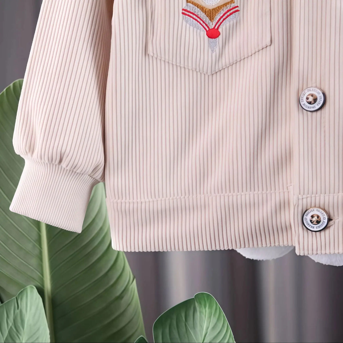A light pink corduroy jacket with a decorative patch is shown on a hanger.