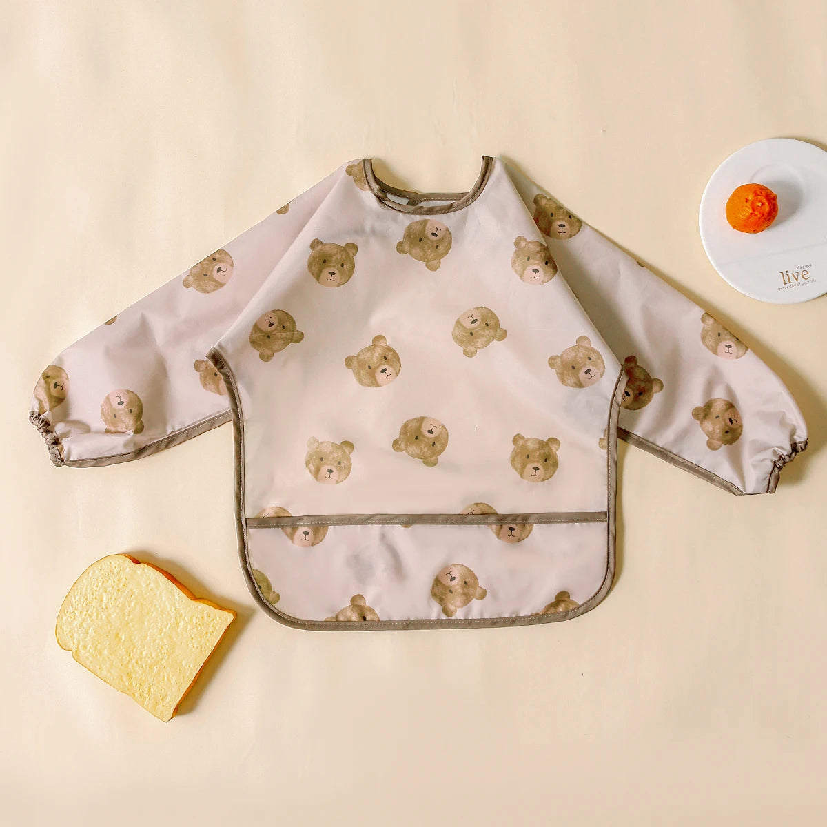 A light pink waterproof long sleeve feeding bib featuring a pattern of small, light brown bear faces for babies.