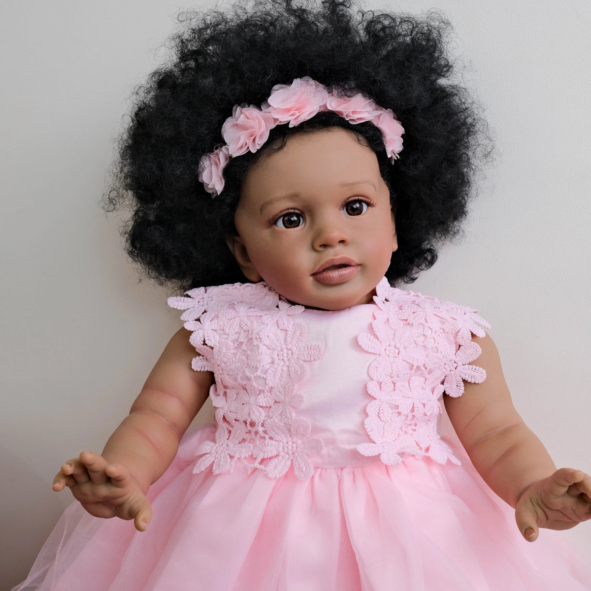 A lifelike reborn toddler doll with hand-rooted black hair wearing a pink dress and floral headband.
