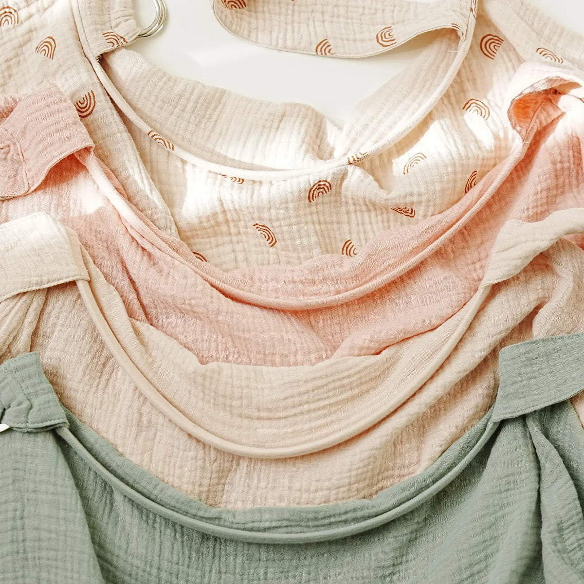 A close-up of a layered set of breathable muslin nursing covers in shades of peach, pink, and sage green.