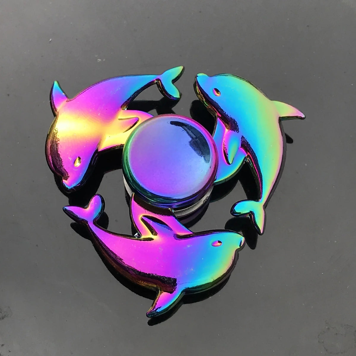 A rainbow metal fidget spinner featuring three stylized fish shapes and a central sphere rotating on a dark surface.
