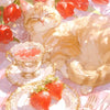 A fluffy orange cat sits next to a cup of tea and a plate of strawberries on a pink checkered surface.