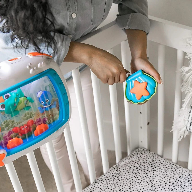 A hand is placing an orange star-shaped toy into the clear viewing window of an ocean-themed crib soother.