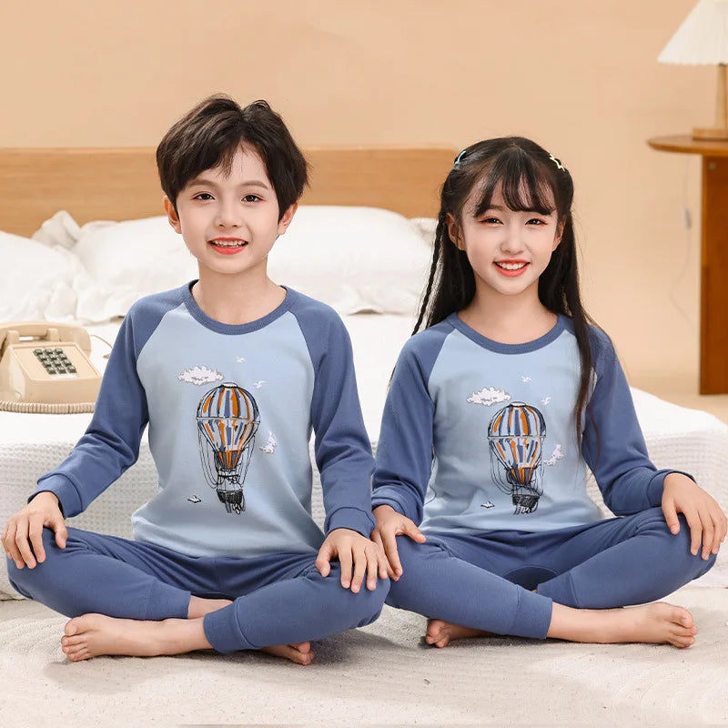 A boy and a girl wearing matching blue cotton pajama sets with an astronaut theme sitting on a bed.