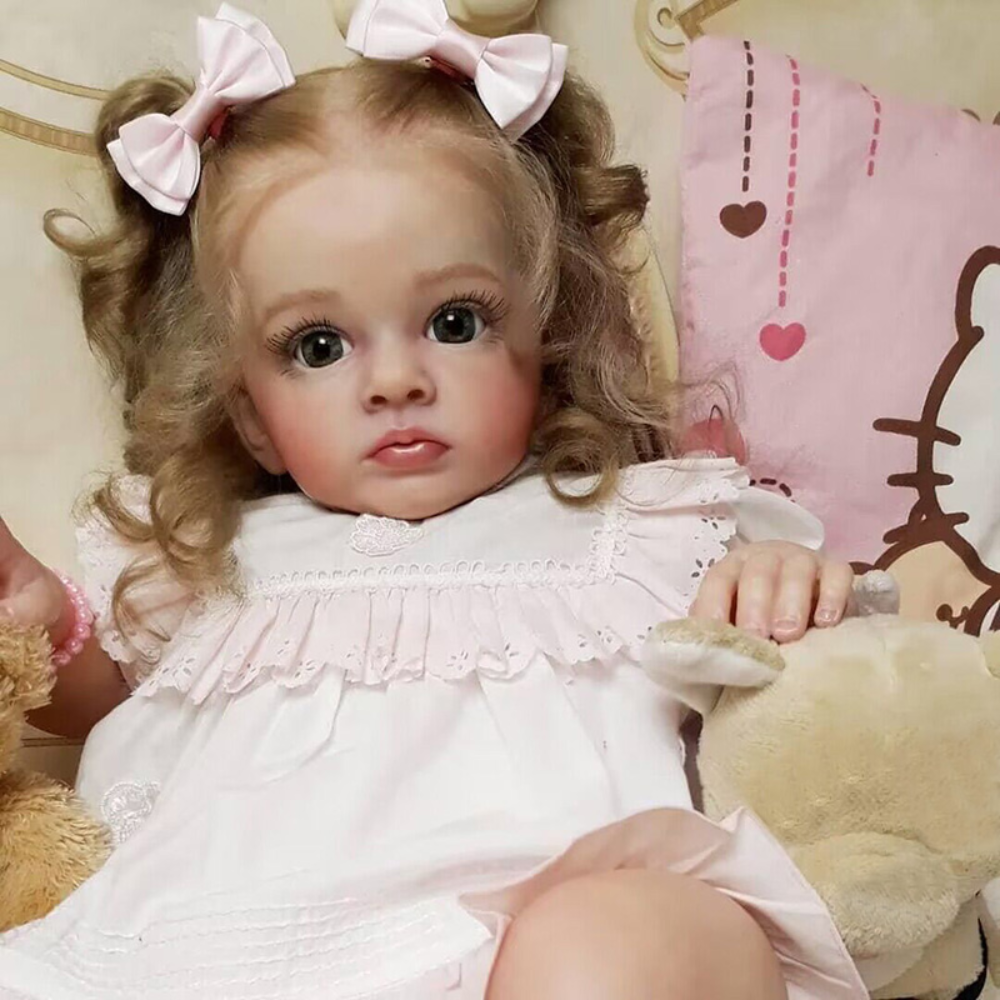 A lifelike reborn baby doll with pink hair and a white dress sitting in a soft, light-colored setting.