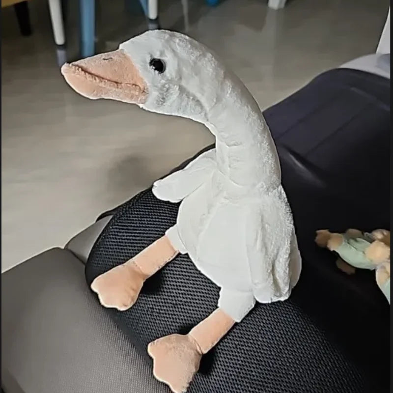 A white long neck goose plush doll with pink feet is resting on a dark gray sofa.
