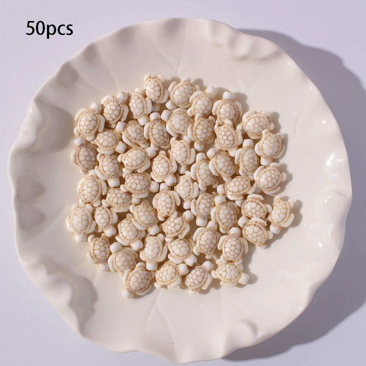 A white plate filled with 50 small, light beige and white ocean turtle beads for jewelry making