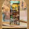 A DIY book nook kit featuring a detailed miniature scene with LED lighting and a dust cover displayed in an arched frame.
