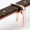 A close-up of a rose gold metal guitar capo with a pick holder mounted on the neck of a wooden guitar fretboard.