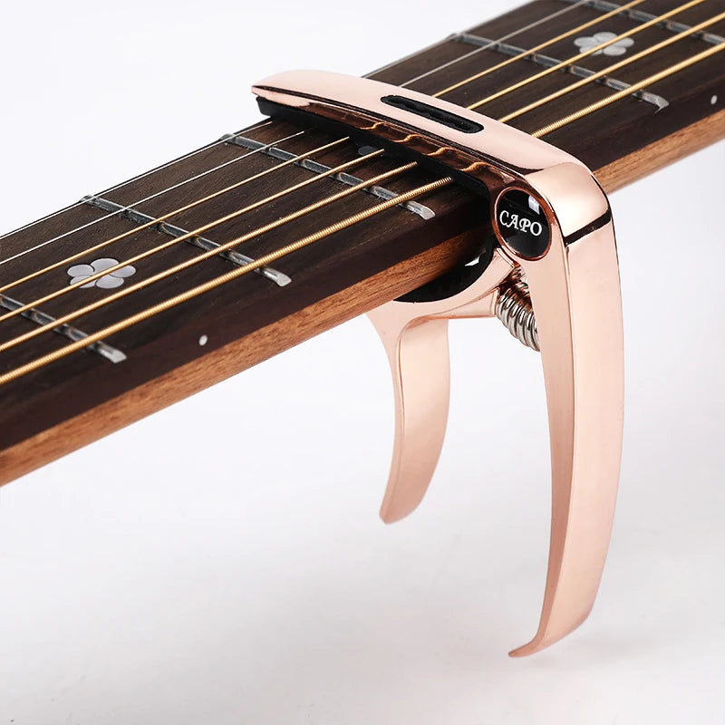 A close-up of a rose gold metal guitar capo with a pick holder mounted on the neck of a wooden guitar fretboard.