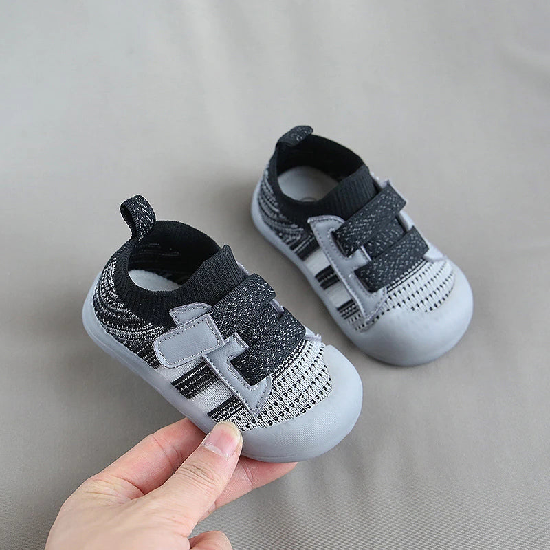 A hand holds a pair of gray and black breathable mesh toddler walking shoes with an anti-slip sole.