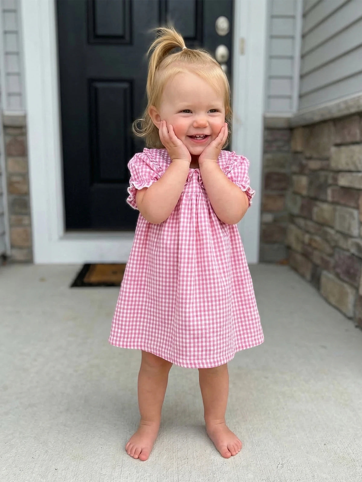 A toddler girl wearing a pink gingham A-line dress with a Peter Pan collar standing outdoors.