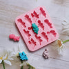 A pink silicone mold with dinosaur shapes is shown on a white wooden surface, surrounded by small colored dinosaurs.