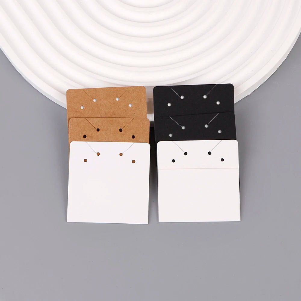A display of square jewelry cards in kraft brown, black, and white, arranged on a gray surface.