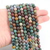 A hand holds a strand of round, loose natural India agate beads in various shades of green, brown, and purple.
