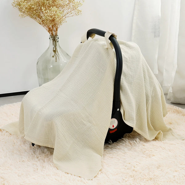 A beige breathable cotton muslin car seat cover draped over a black carrier on a fluffy rug.
