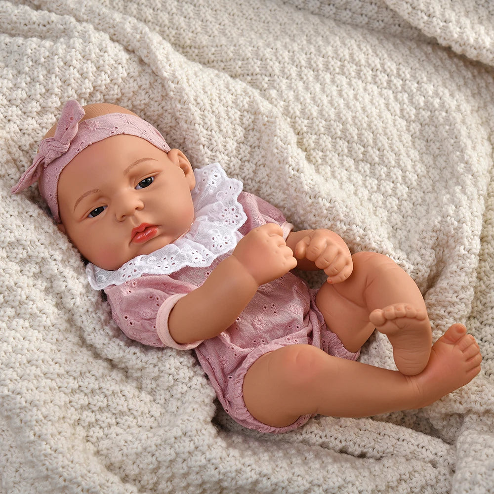 A lifelike vinyl reborn doll with a pink outfit and headband lying on a white knitted blanket.