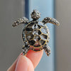 A detailed rhinestone turtle brooch pin with a textured shell and gold accents is held by a hand.