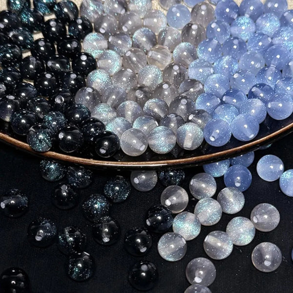 A close-up of various iridescent and translucent resin beads in shades of black, gray, and blue scattered on a dark surface.