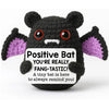 A handmade knitted bat doll with black body, white mouth, and purple ears, featuring a positive message.