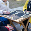 Two people play a game of double-six dominoes on a wooden table outdoors.
