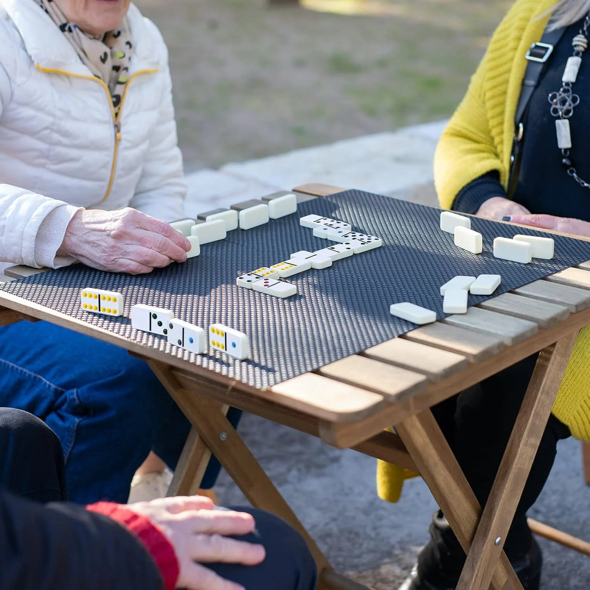 Two people play a game of double-six dominoes on a wooden table outdoors.