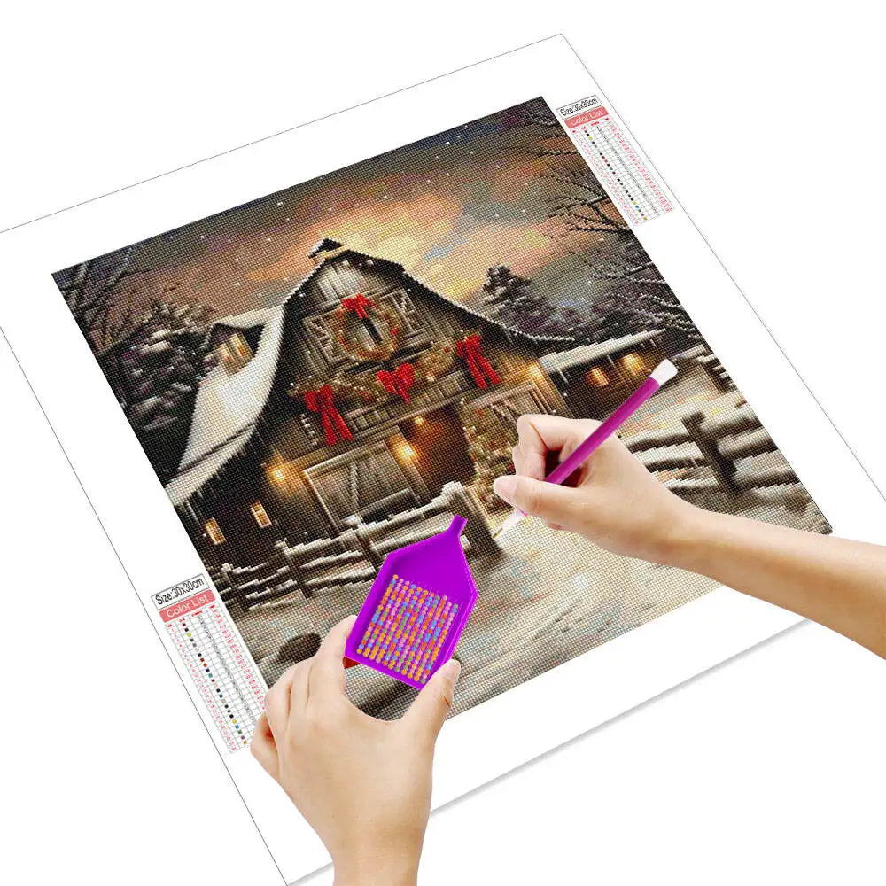 Hands are painting a detailed Christmas village scene on diamond painting canvas with a drill and colored beads.