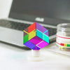 A colorful CMY prism cube with a hexagonal shape resting on a white stand next to a laptop.