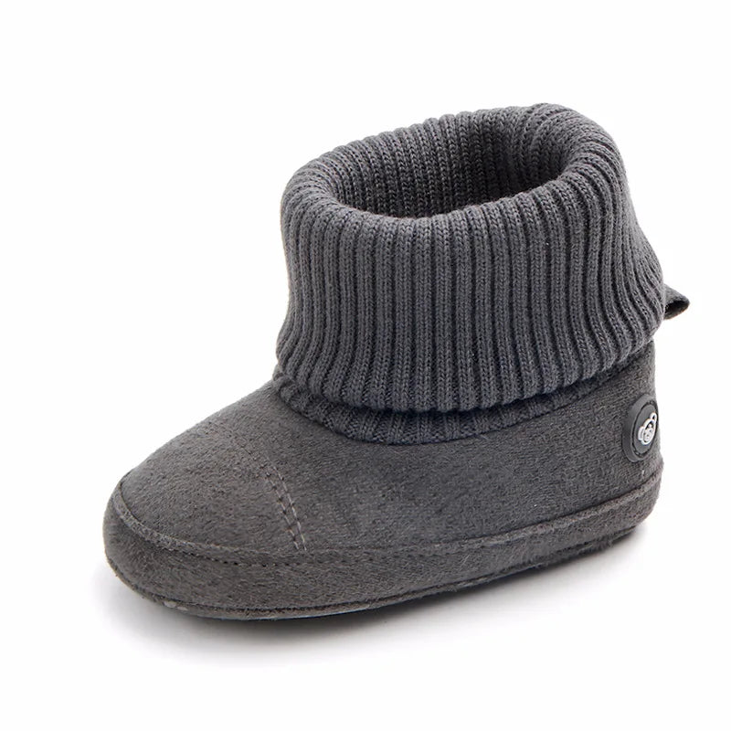 A pair of gray knitted baby winter snow boots with a ribbed cuff and fuzzy ball accents.