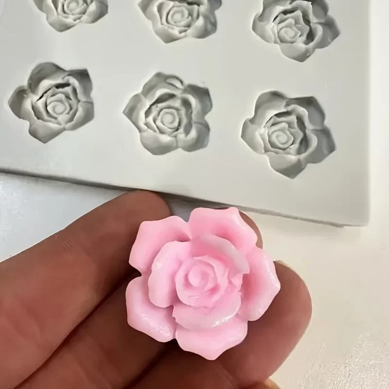 A hand holds a pink rose flower silicone mold next to a white silicone tray with gray rose molds.
