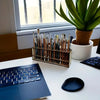 A wooden art brush and pencil organizer with 67 slots sits on a white desk next to a laptop and a potted plant.