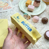 A yellow butter stick squishy toy is held in a hand, surrounded by various cosmetic and fidget items.
