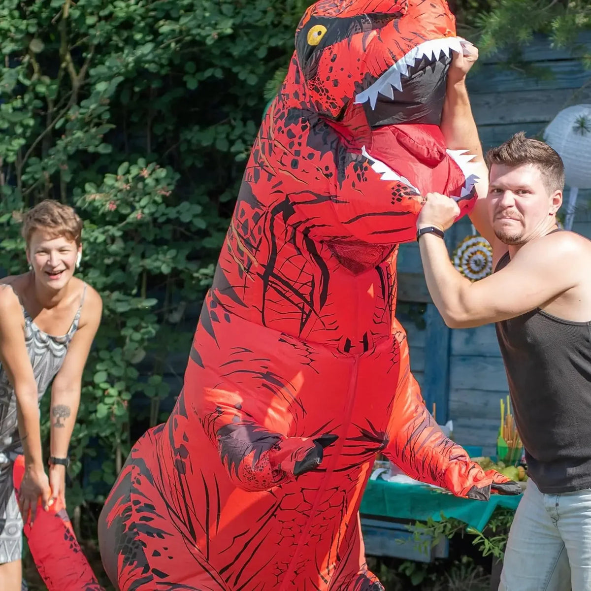 A man is posing with an inflatable red and black T-Rex costume in an outdoor garden setting.
