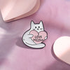 A white cat enamel pin with a pink heart design is placed on a soft pink surface.