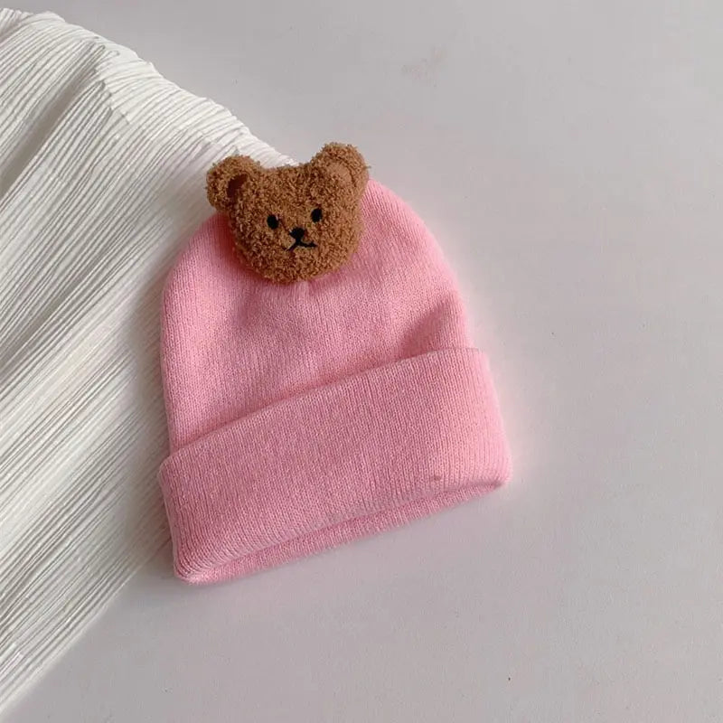 A pink knitted baby hat with a brown bear ear decoration resting on a white ribbed fabric.