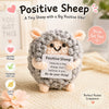 A handmade gray crochet sheep doll with a motivational card and pink flower on a beige fabric.