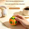A hand holds an orange and green 3D gear ball fidget toy on a light surface next to headphones and a notebook.